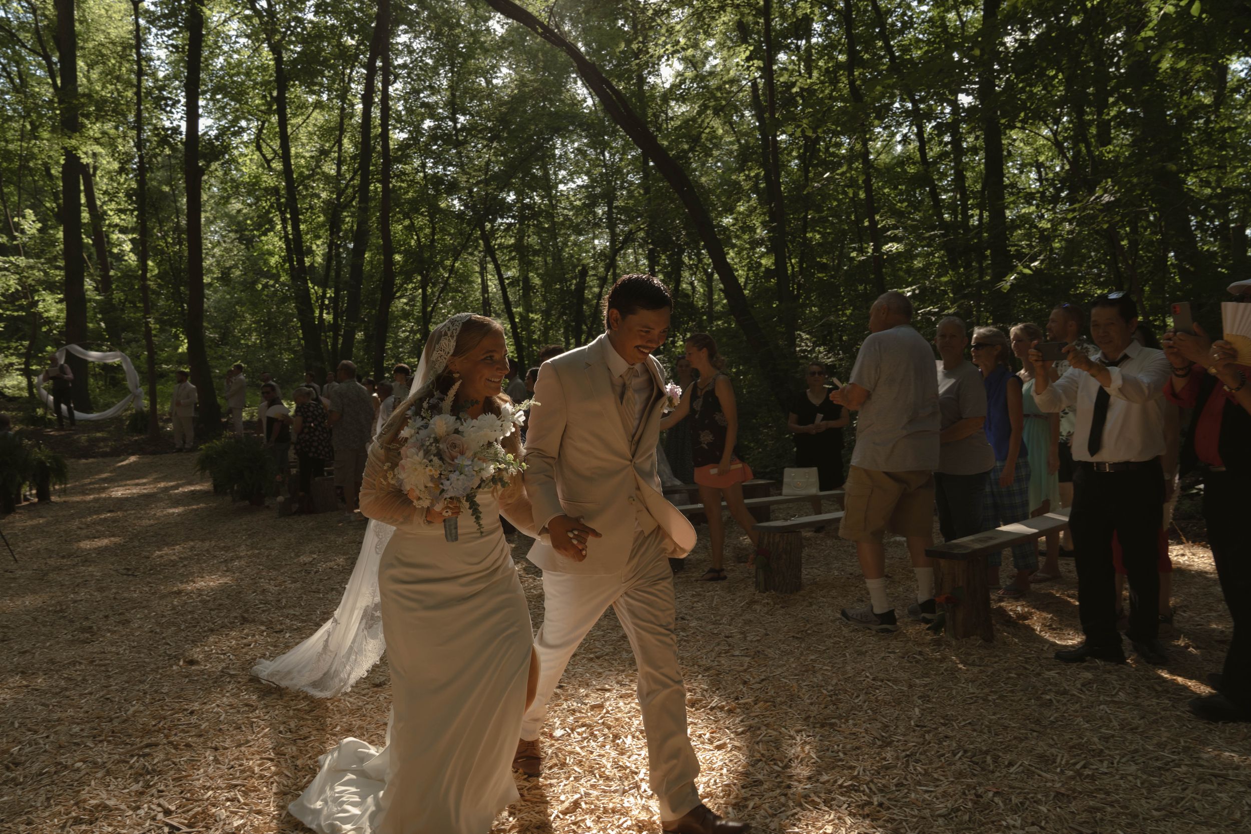 A couple celebrates their wedding ceremony in a forest setting surrounded by trees.