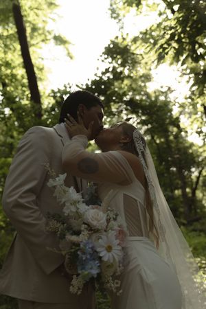 A couple shares their first kiss as newlyweds in a forest with white floral bouquet.