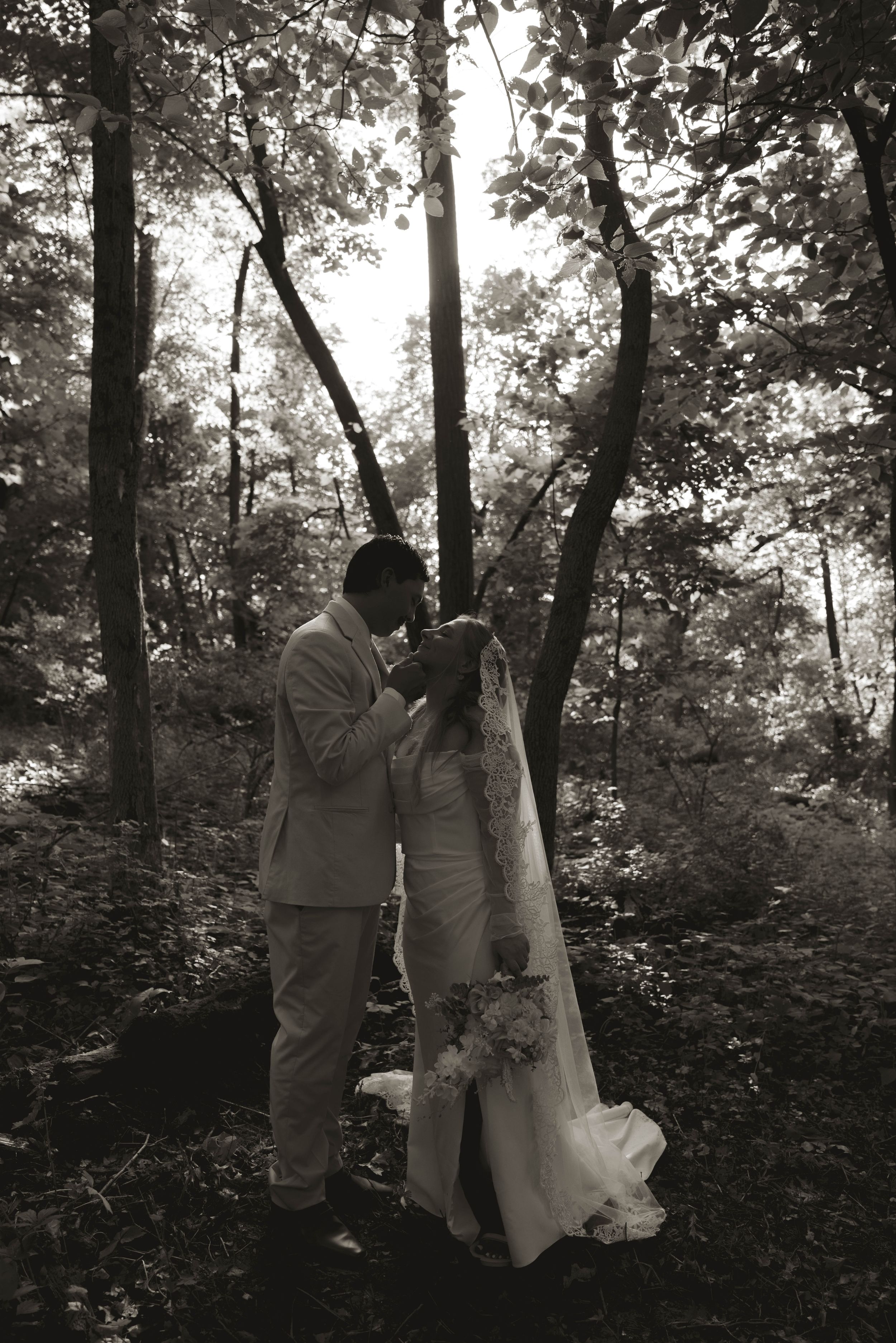 A couple shares an intimate moment in a wooded setting wearing light colored formal attire.