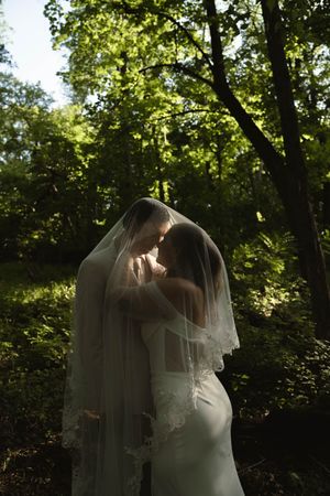 A bride in a flowing white dress and veil walks through a sunlit wooded area during a wedding photoshoot.