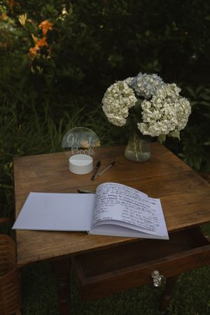 A wedding guestbook with white flowers lies open on a rustic wooden table outdoors.