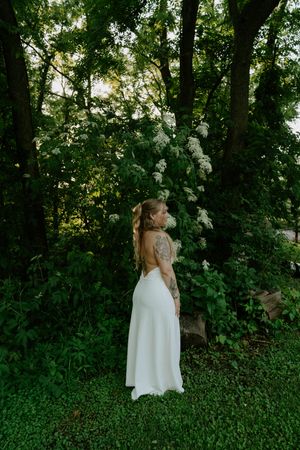 A white wedding dress stands out against lush greenery and flowering bushes in a forest setting.