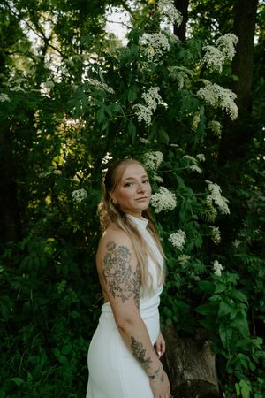 An elegant white wedding gown is showcased among blooming white flowers in a wooded setting.
