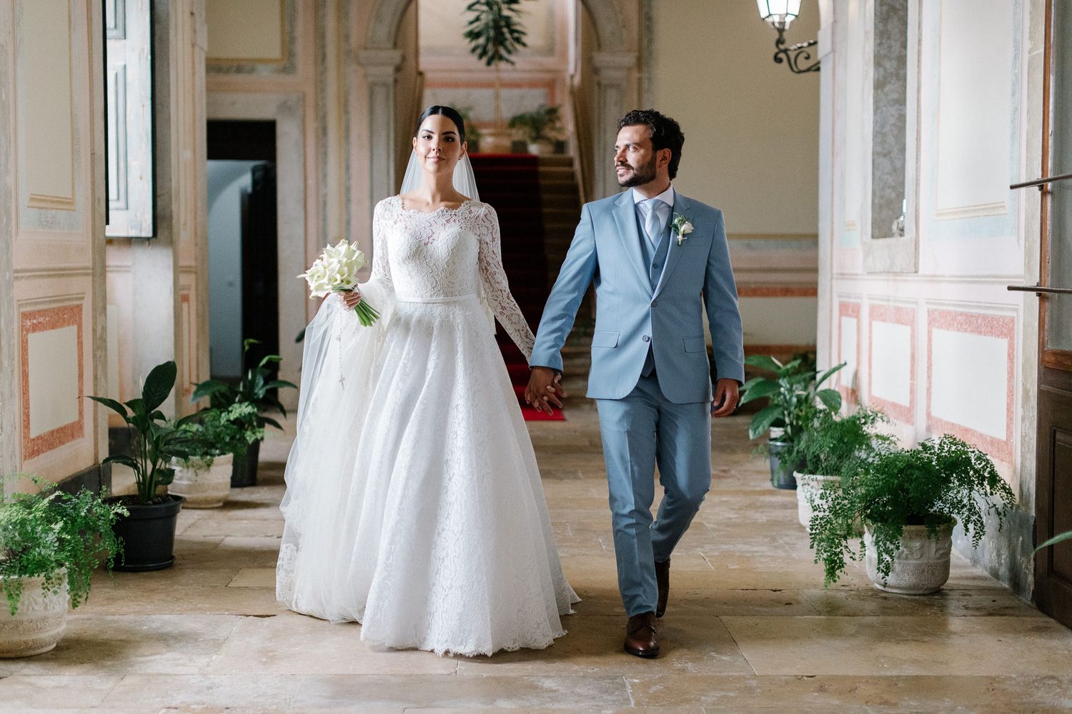 A couple in wedding attire walks down an elegant hallway with ornate walls and traditional lantern lighting.