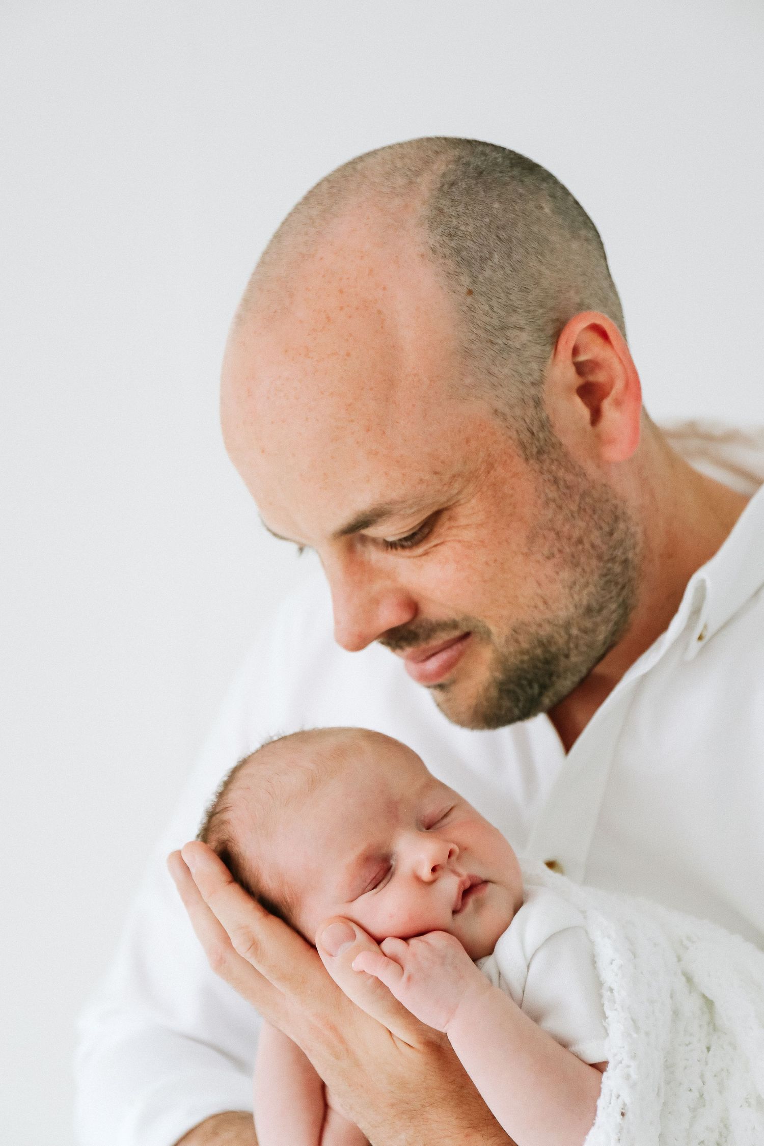 Dad with newborn being held and gently kissed while wearing white against a white background, during newborn photoshoot in Nottingham