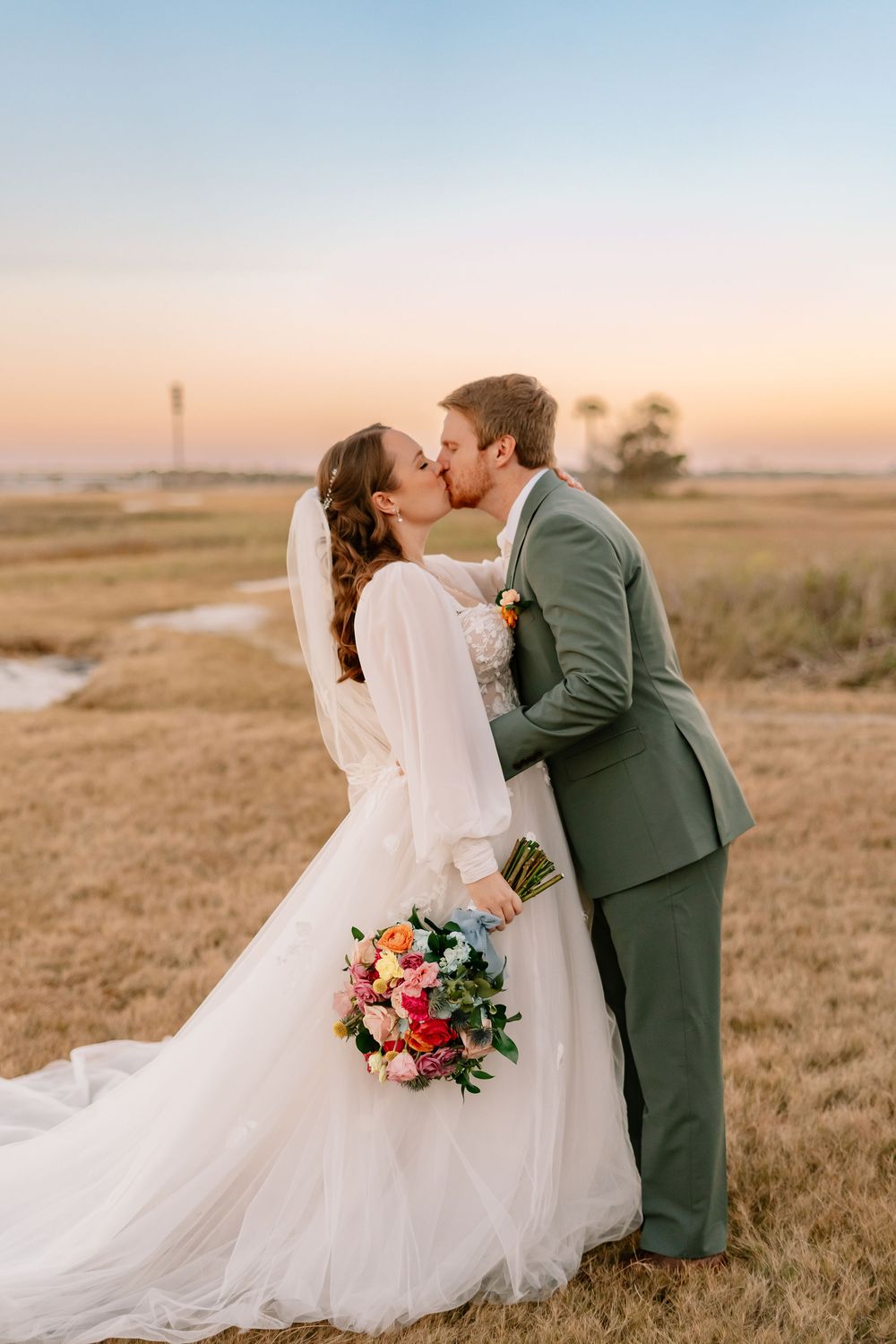 A romantic wedding photo during sunset showing a couple embracing in a field with a colorful bouquet against a golden sky.