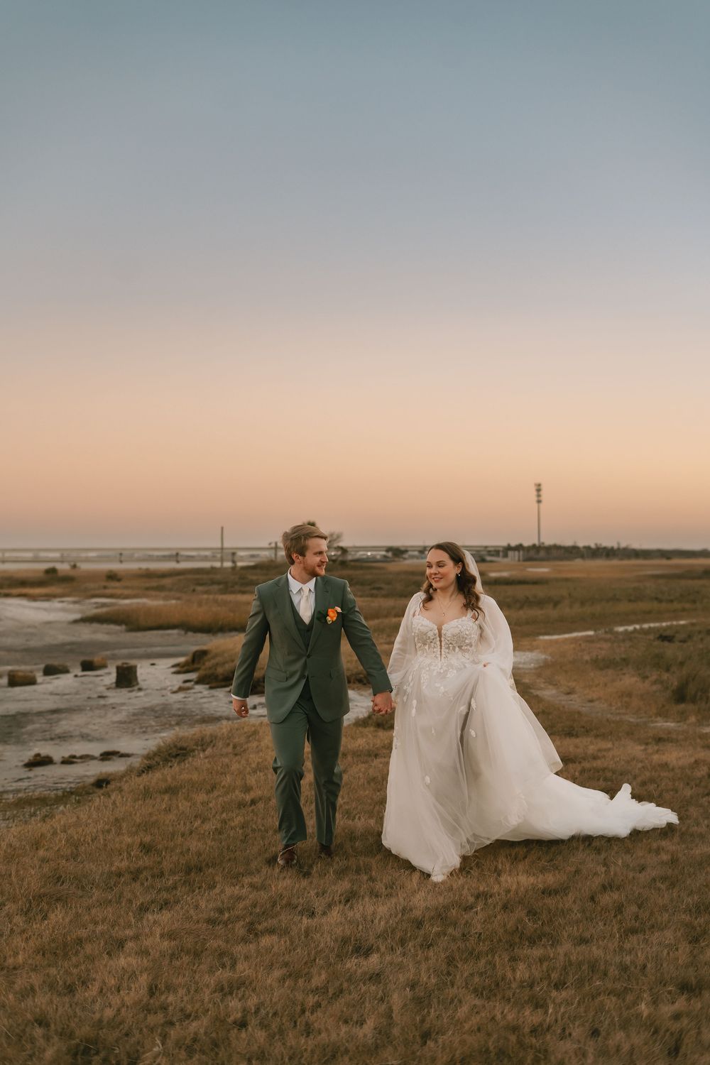 Couple in wedding attire walking through coastal marshlands at sunset with golden light illuminating the landscape.