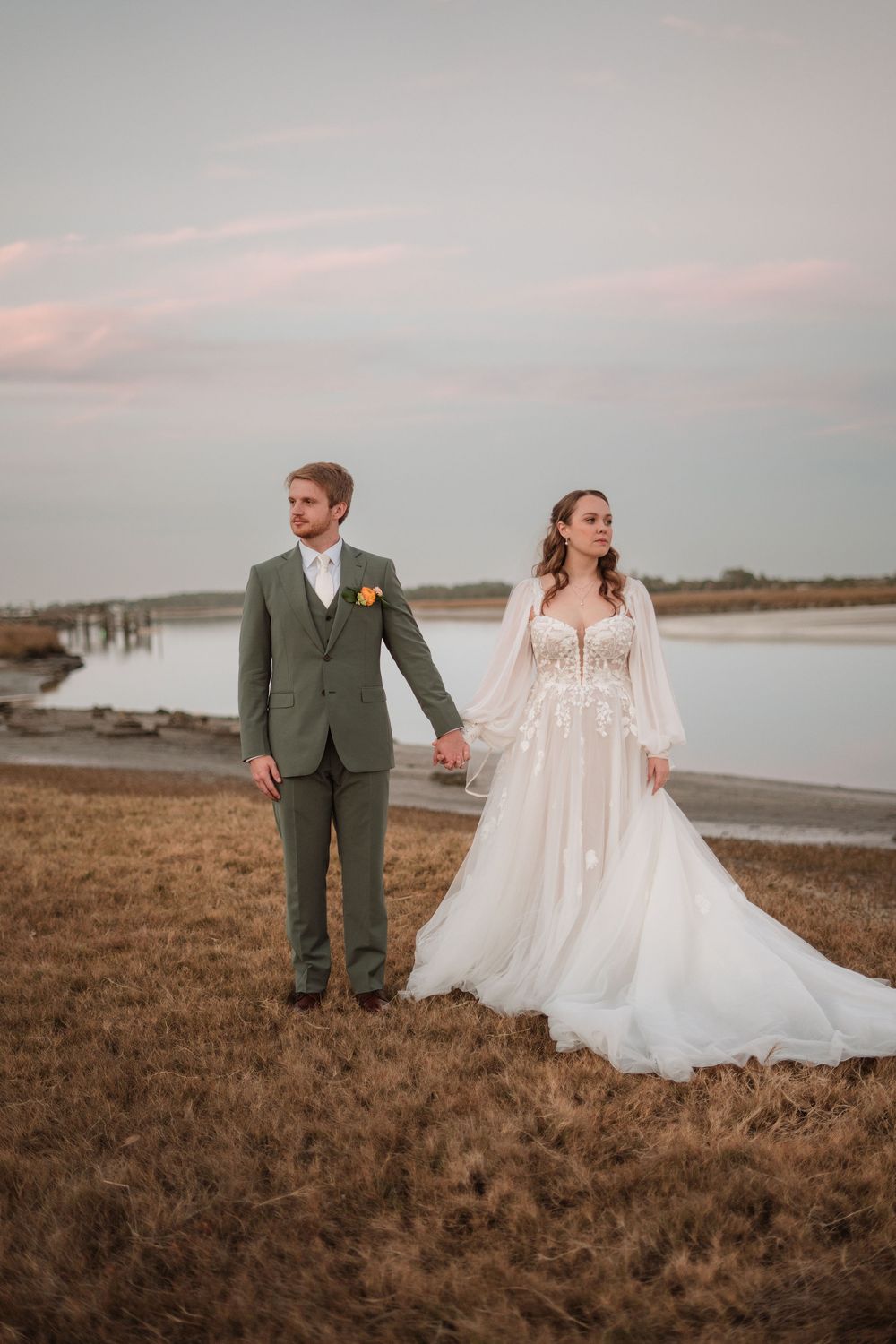 Newlywed couple walks along grassy coastal landscape at sunset in romantic wedding photo series.