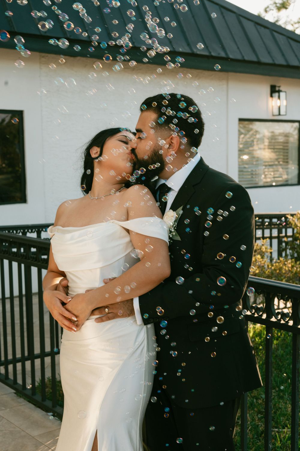A couple embraces in a tender kiss surrounded by floating bubbles near a white building.