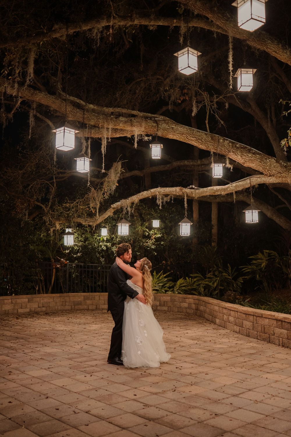 Outdoor wedding dance underneath glowing lanterns hung from tree branches on a stone patio at night.
