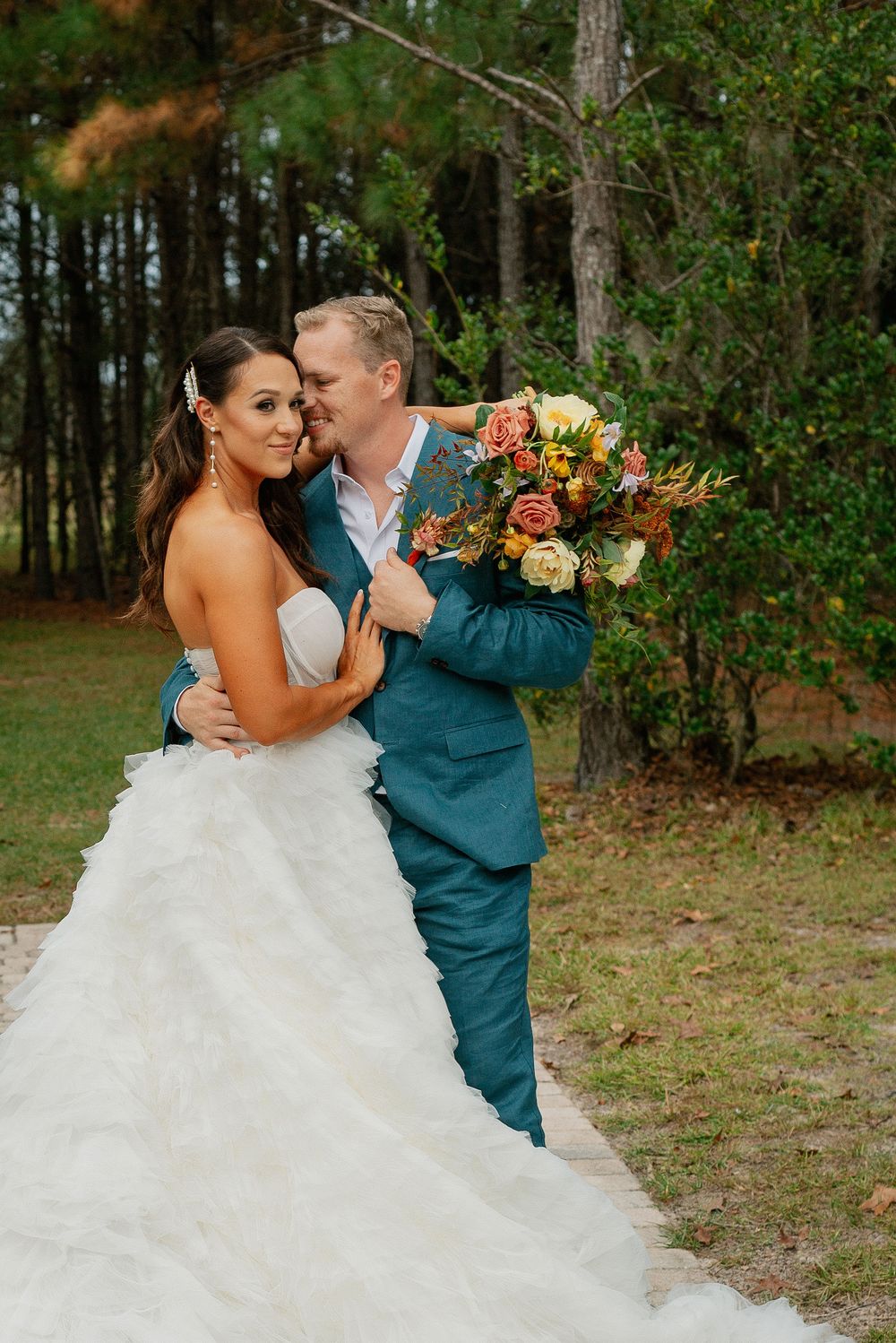 A romantic outdoor wedding photo of a couple embracing, with the bride in white and groom in teal holding autumn flowers.