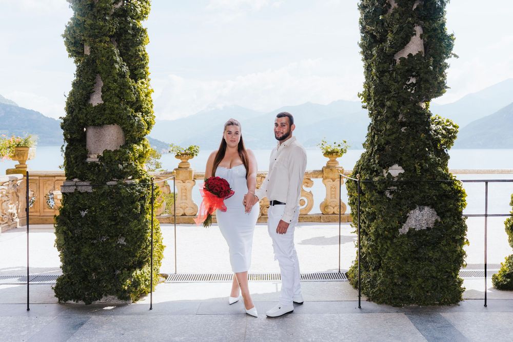 A wedding couple walks along a scenic lakeside venue with mountains and greenery in the background.