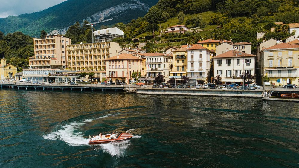 A speedboat races across blue waters along a coastal town with mountains and charming waterfront buildings in Italy.