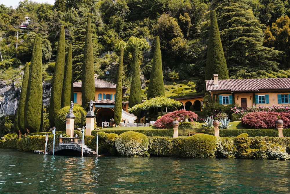 Luxurious lakeside villa with tall cypress trees and colorful gardens along Lake Como's shoreline in Italy.