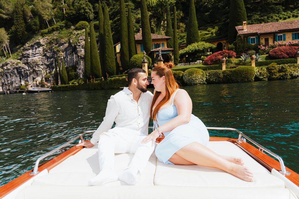Couple lounges on wooden boat at Lake Como with scenic waterfront villas and cypress trees in background.