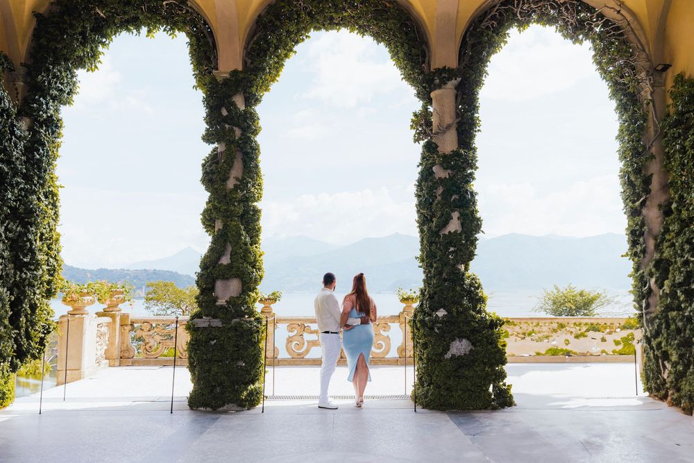 Romantic vine-covered archway frames a coastal view with a couple walking along the Mediterranean Sea.