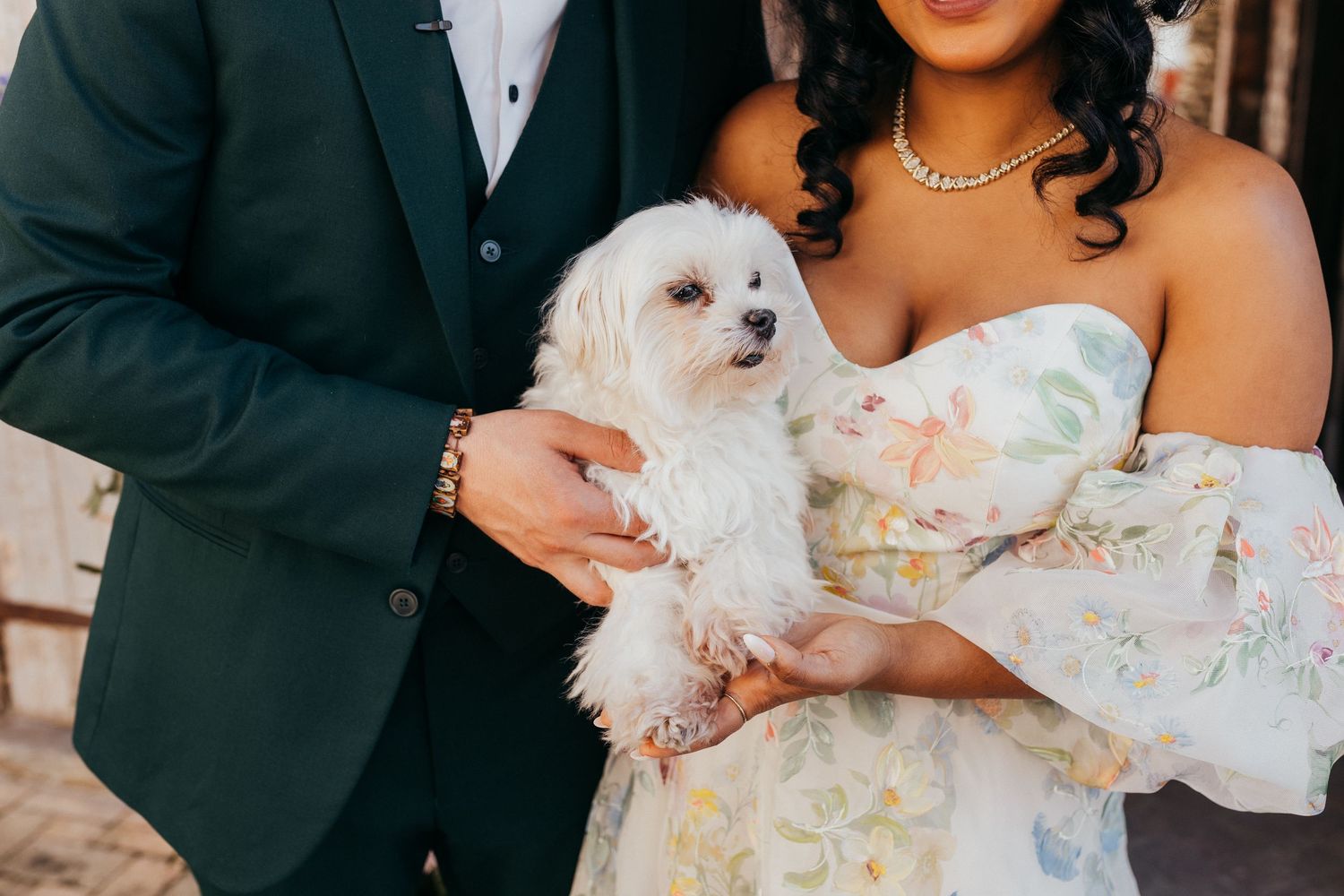A couple holds their small white Maltese dog while wearing formal wedding attire including a green suit and floral dress.