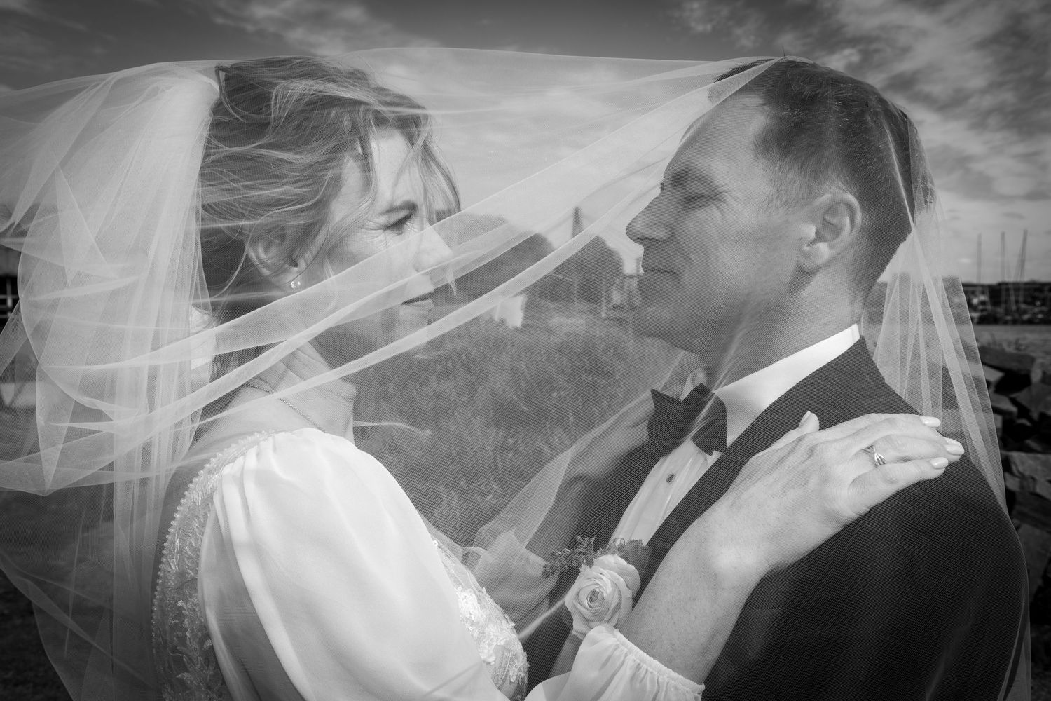 Romantic black and white photo of bride and groom under flowing veil with cloudy sky backdrop.