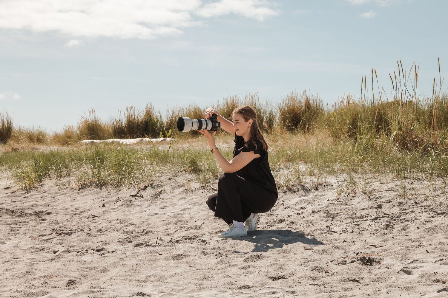 A figure in a black dress crouches down to take a photo on a sandy beach with tall grass in the background.
