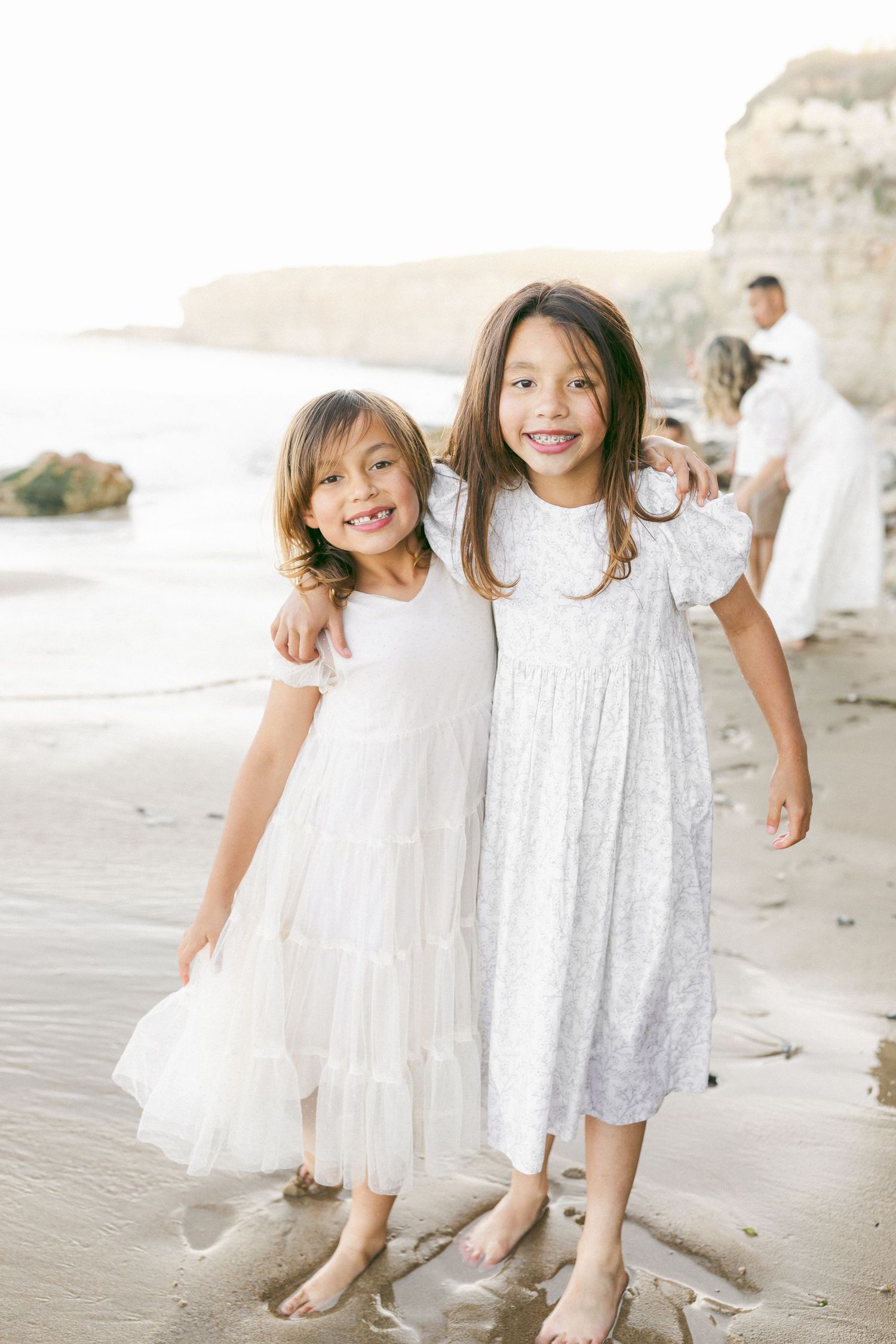 Two young children in white dresses playing and laughing together on a beach with waves in the background captured by Chelsee Rawe Photogra.