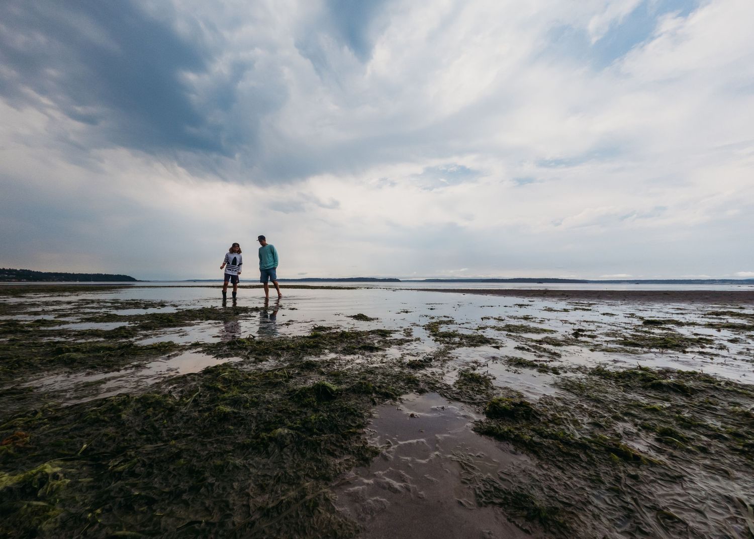 Two silhouetted figures stand on rocky beach at low tide with dramatic cloudy sky overhead.