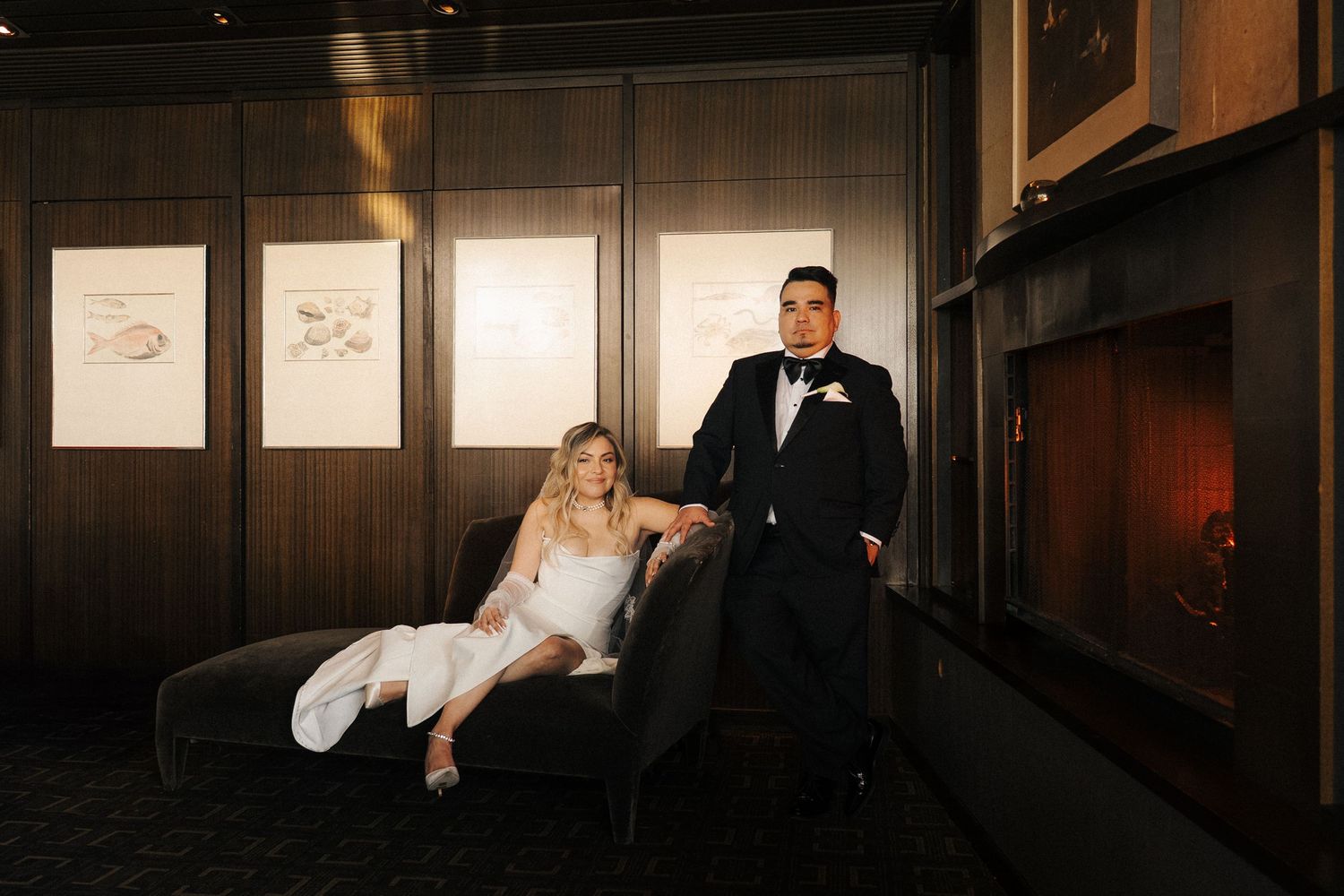 A bride and groom pose elegantly in a dark room with wood paneling and framed artwork on the walls.