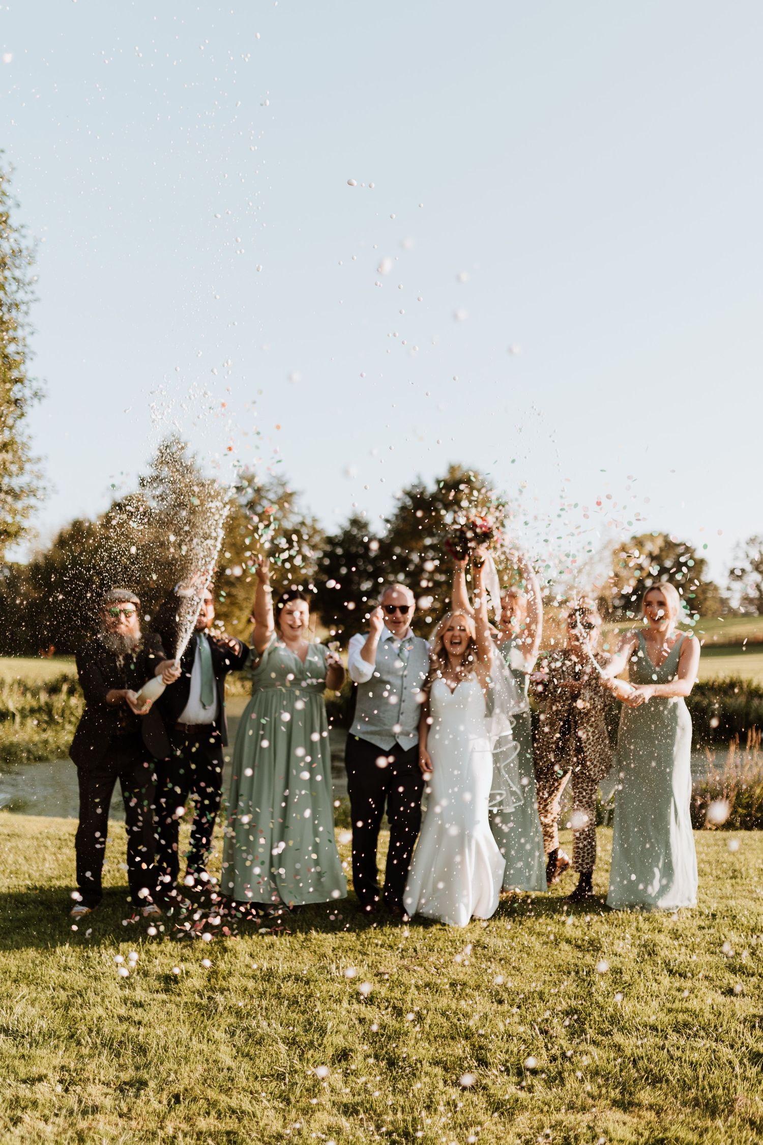 Wedding party celebrates in a sunny field by tossing confetti & spraying champagne in the air during golden hour.