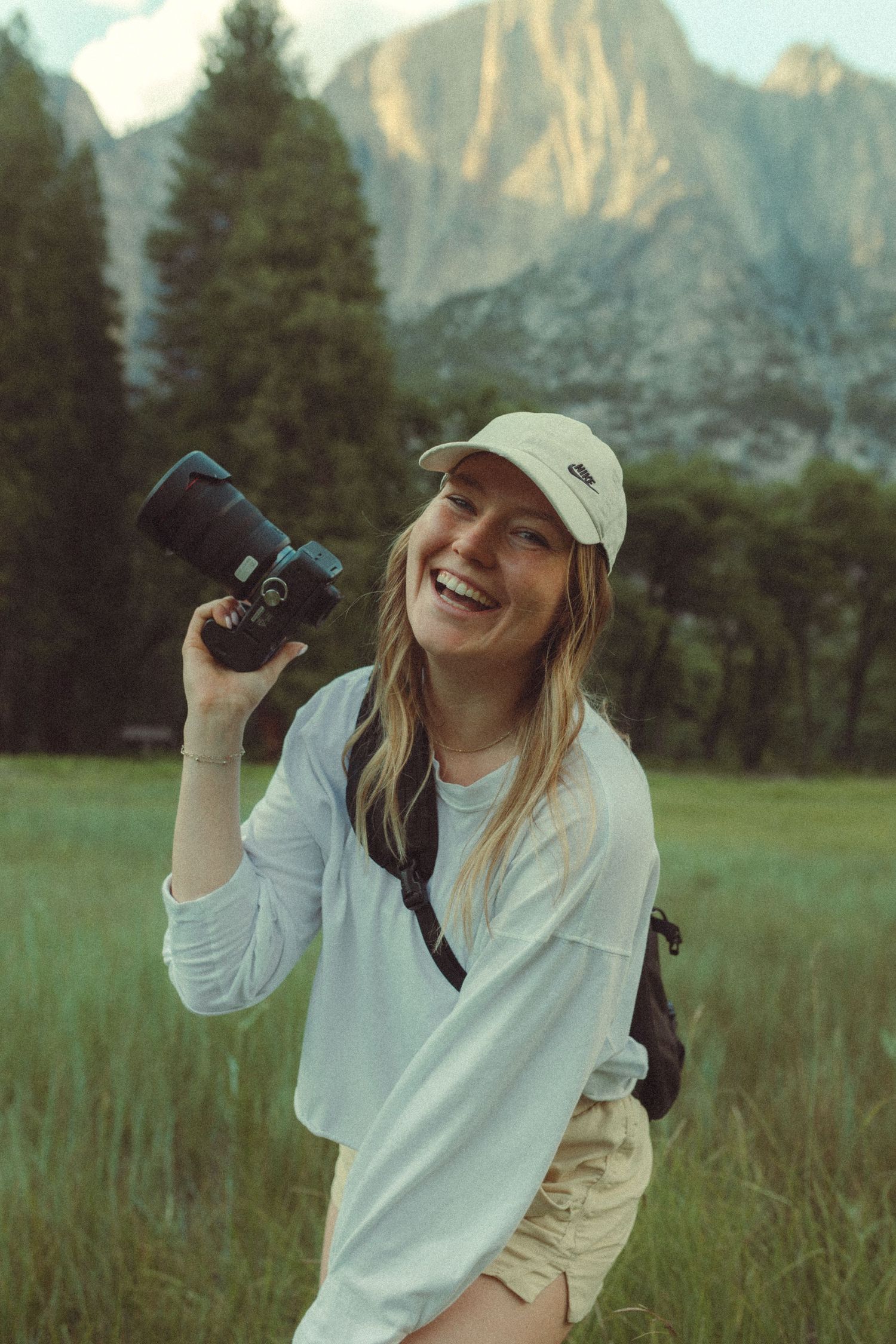 Someone wearing a white sweater and baseball cap uses binoculars to birdwatch in a grassy meadow with mountains behind.