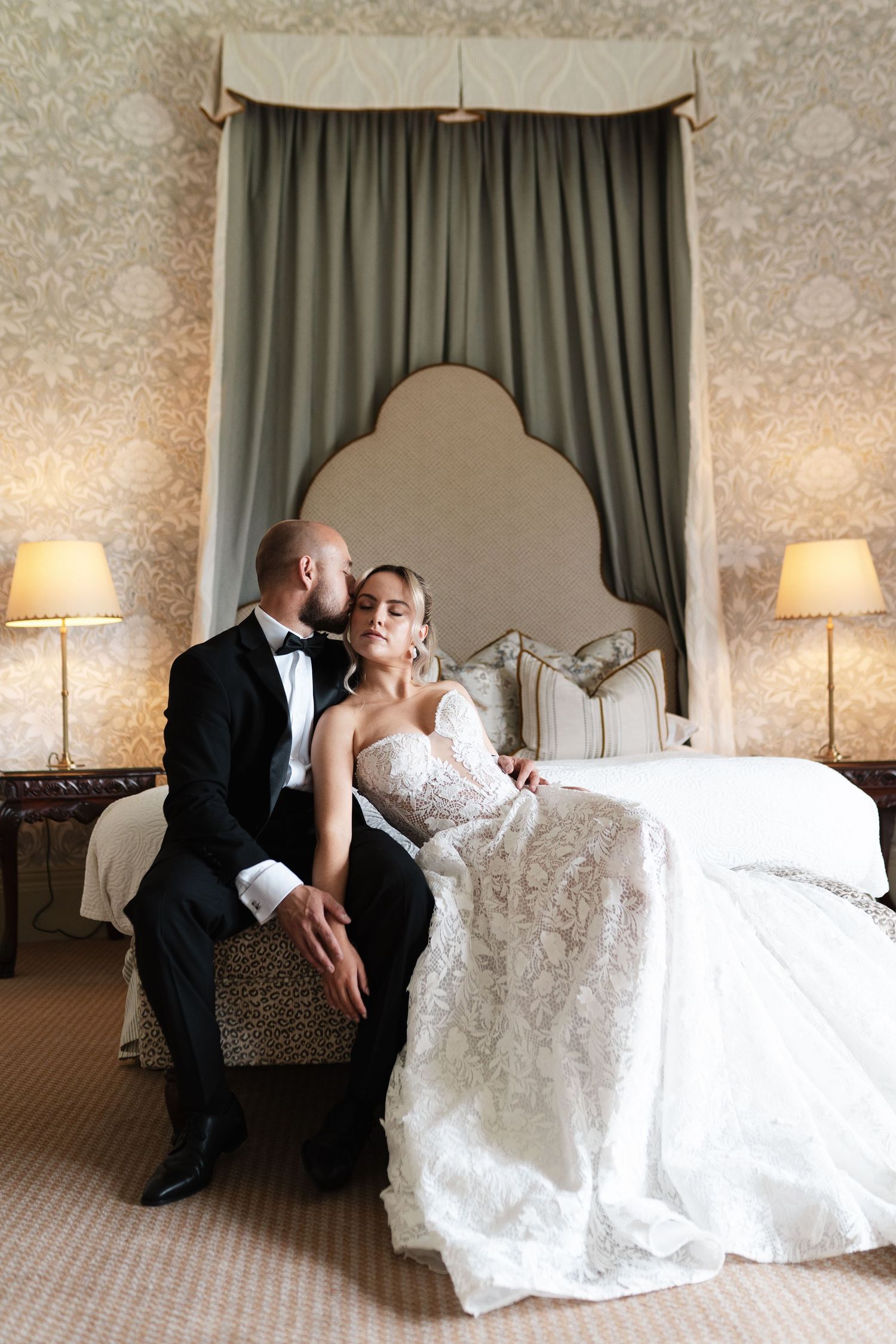 Elegant couple in formal wedding attire share intimate moment while sitting on chair in luxurious bedroom setting.