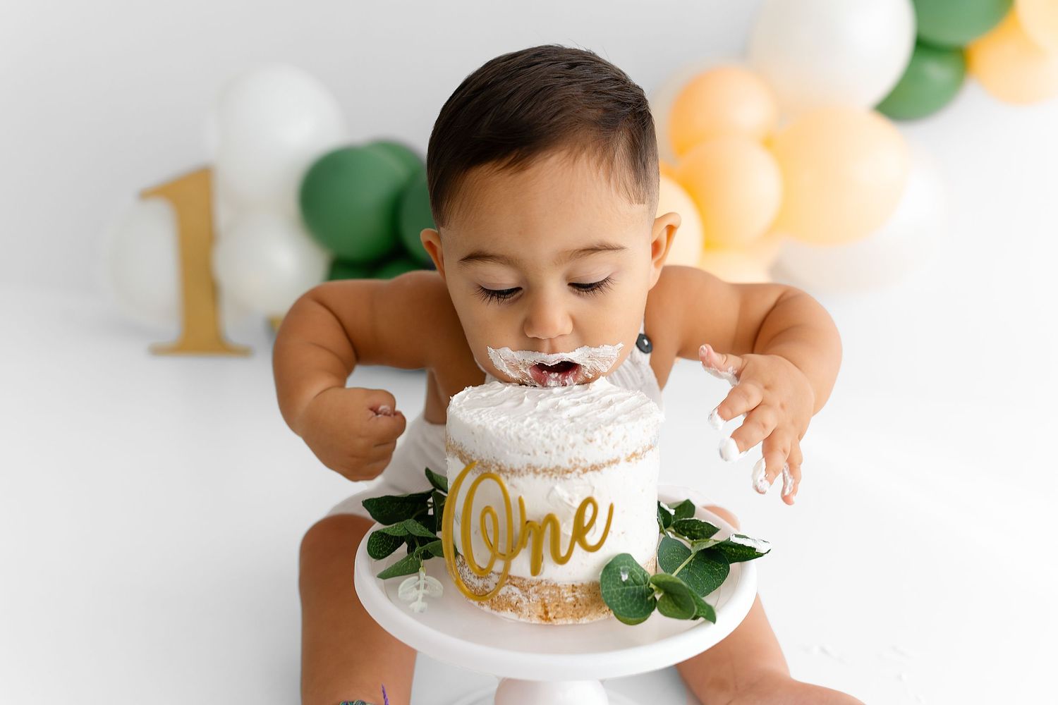 Baby in white celebrates first birthday with smash cake decorated with gold 'One' text and greenery on white backdrop.