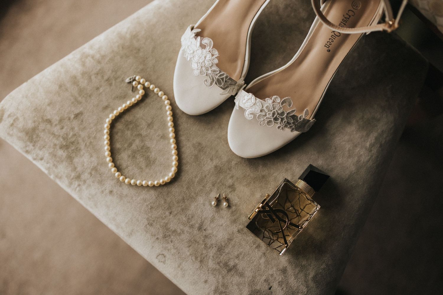 Elegant white bridal heels with pearls and jewelry arranged on textured beige surface for wedding photography.