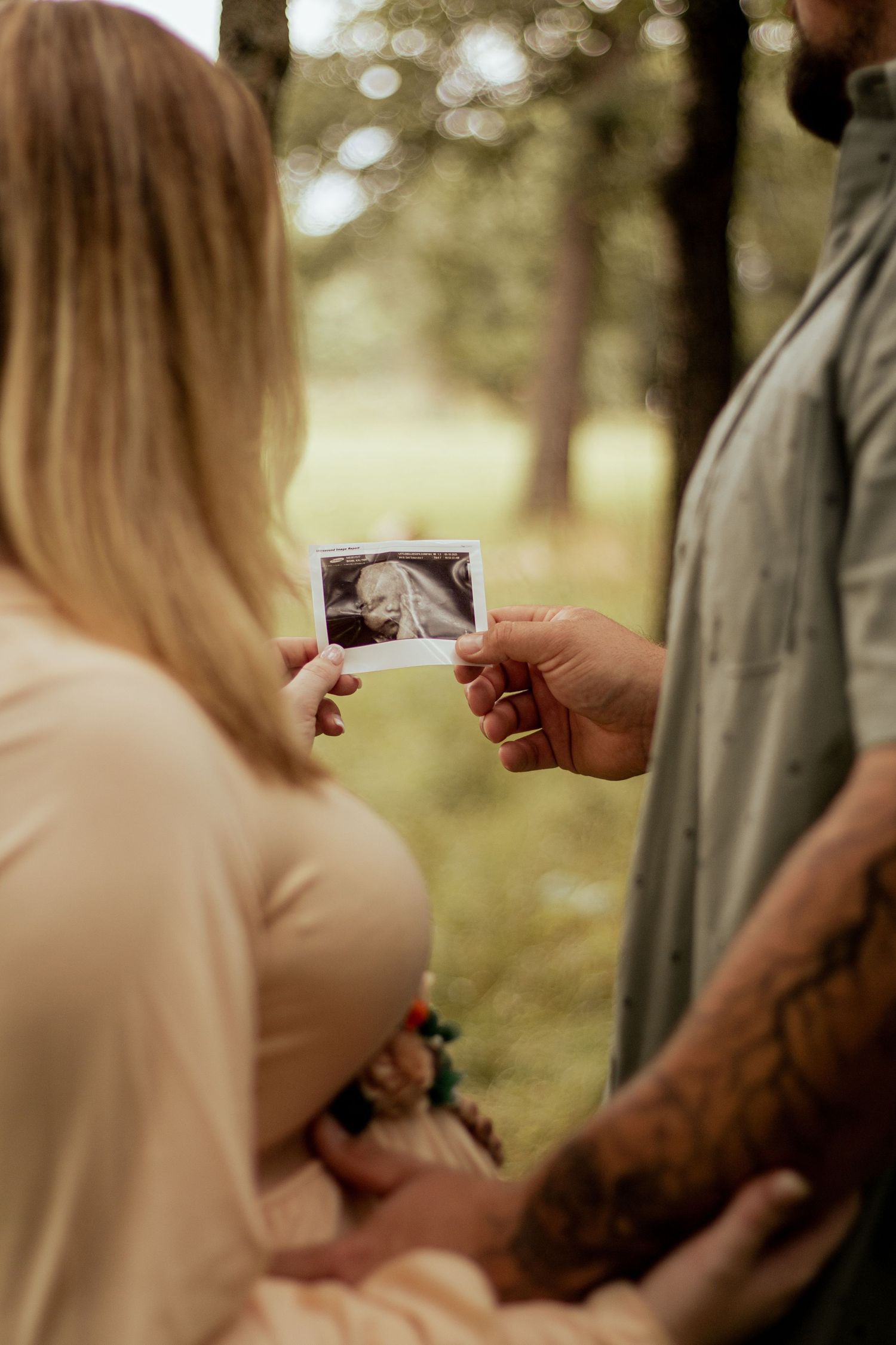 Two people stand in a sunlit wooded area examining a polaroid photograph together on a warm day.