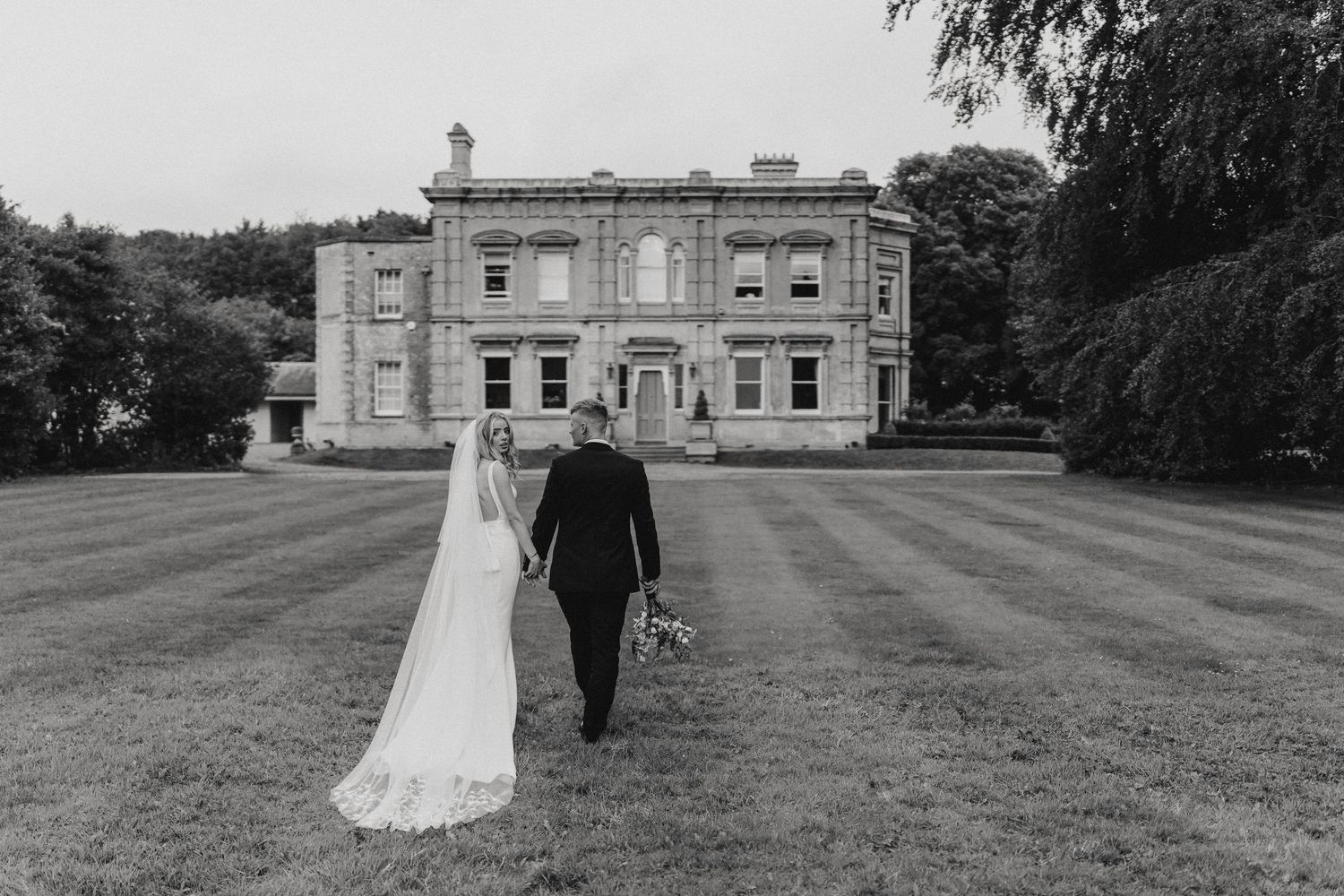 Elegant couple walks on rolling green lawn in front of Cleatham Hall.