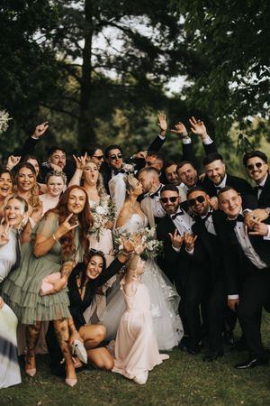 Wedding party in sage green dresses and dark suits pose candidly together on grass making silly expressions and poses.