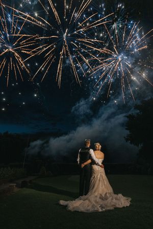 A couple in formal attire watches fireworks burst in the night sky during their outdoor wedding celebration.