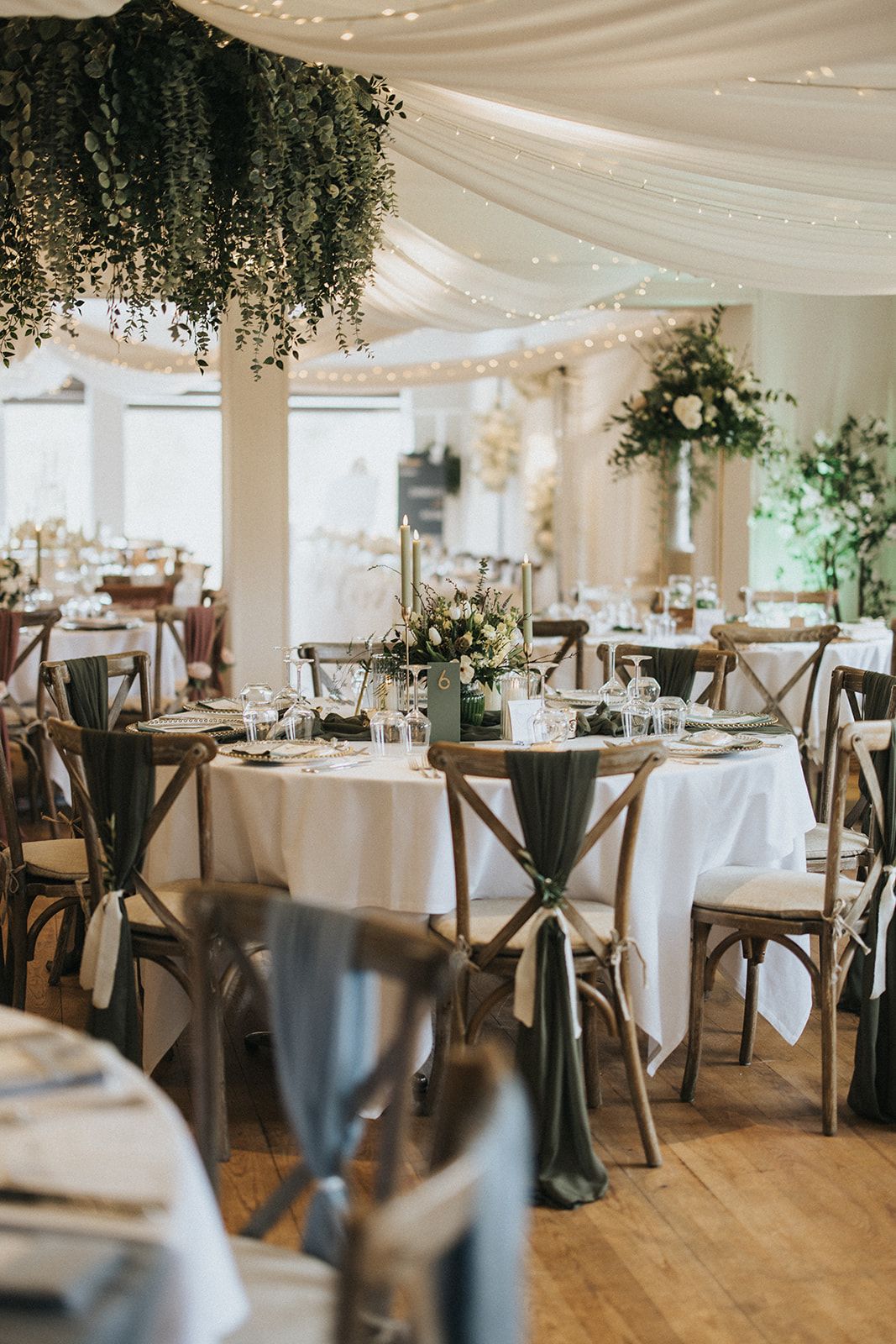Elegant wedding reception venue with white draping, hanging greenery, rustic chairs and round tables set for dining.