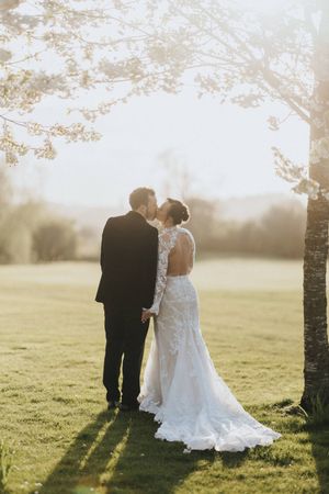 A romantic outdoor wedding photo at sunset with a couple standing among trees in an open field.