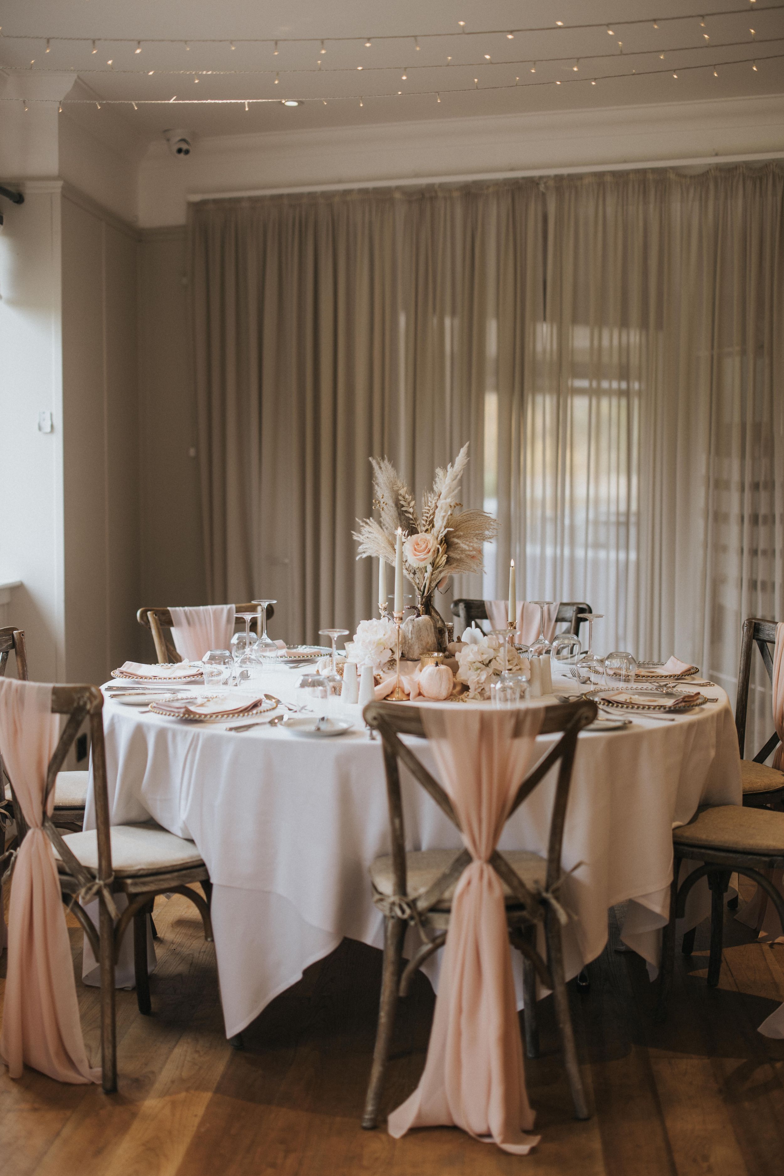 Elegant wedding reception table with white linens, cross-back chairs, and a pampas grass centerpiece under string lights.