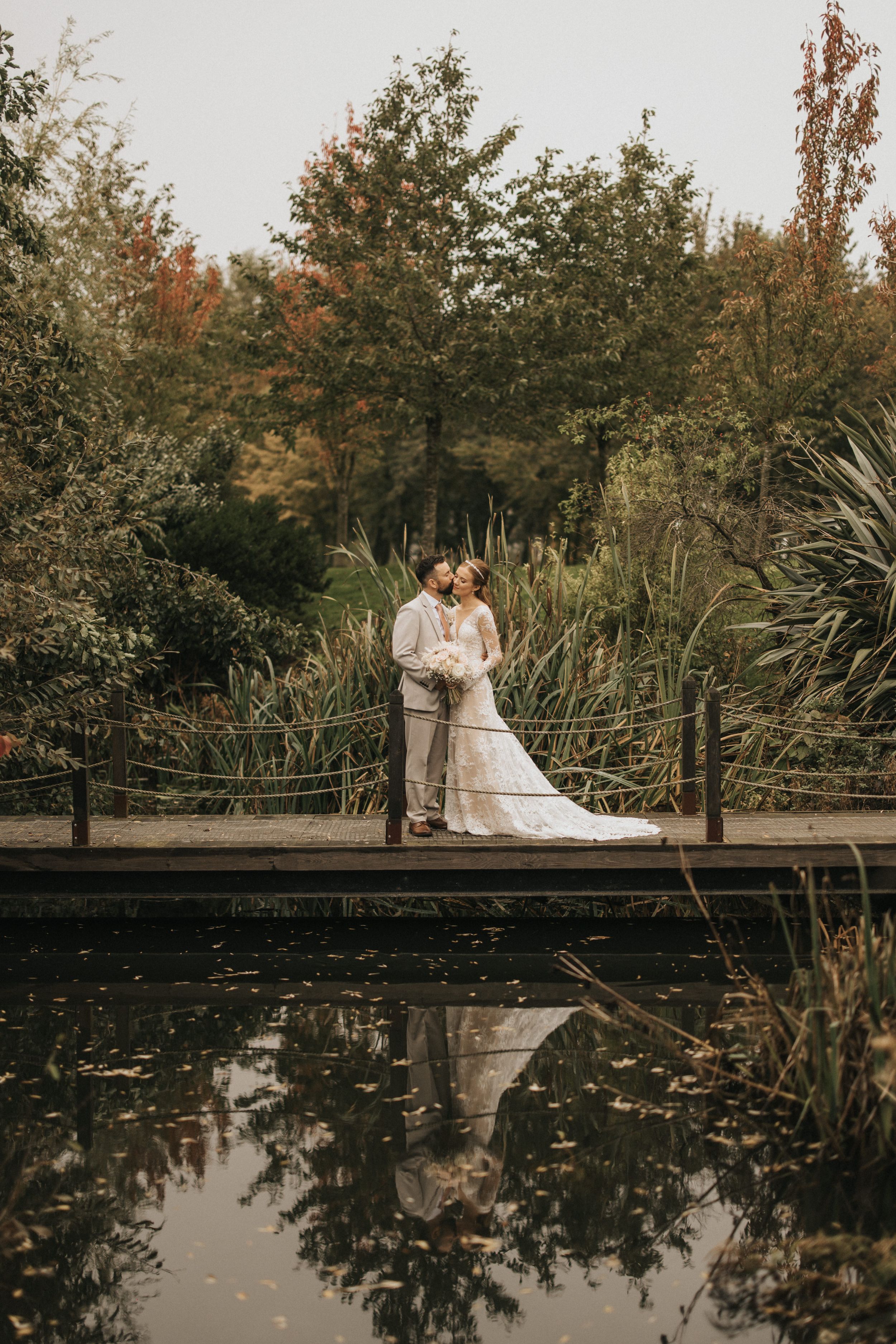 A romantic fall wedding photo taken on a wooden bridge over a pond with reflections amid autumn foliage.