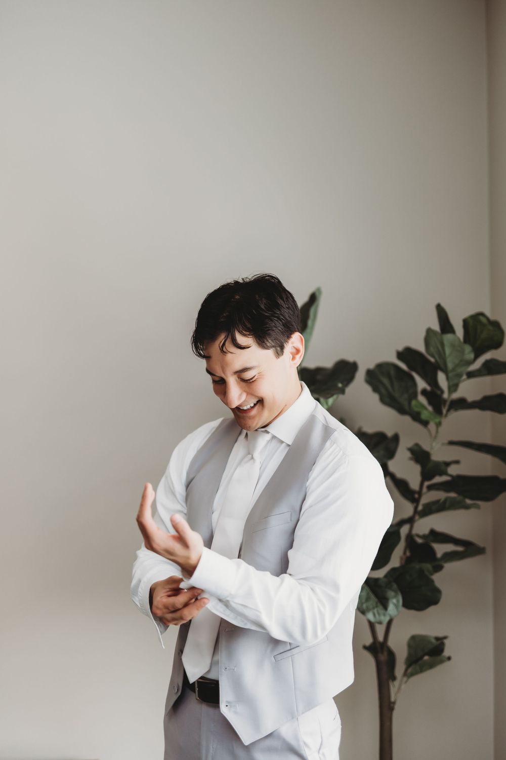 Someone in a crisp white shirt and grey vest adjusts their cuff while smiling and standing next to a potted plant.