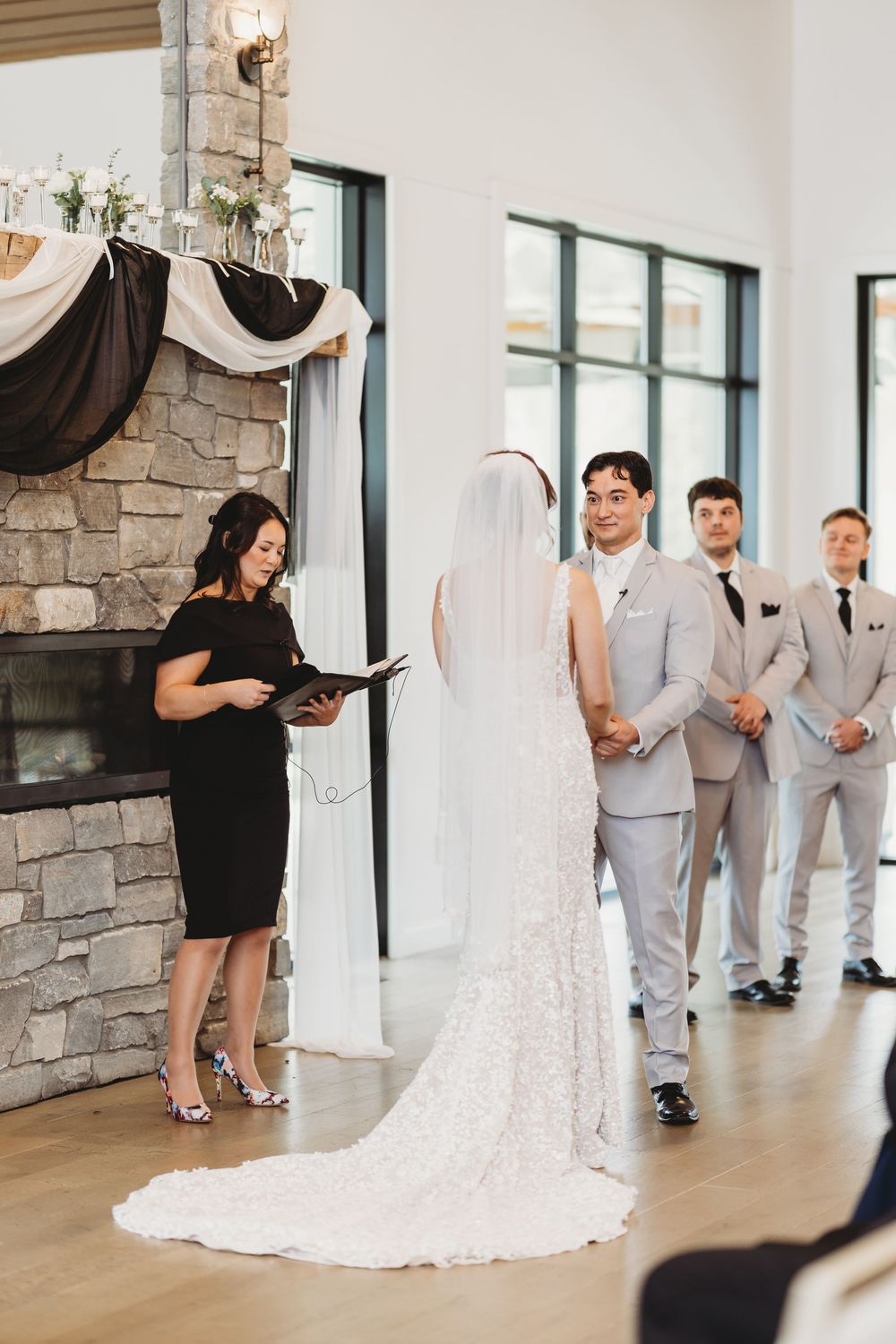 Wedding ceremony in a modern venue with stone fireplace features an officiant in black dress and wedding party in light gray suits.