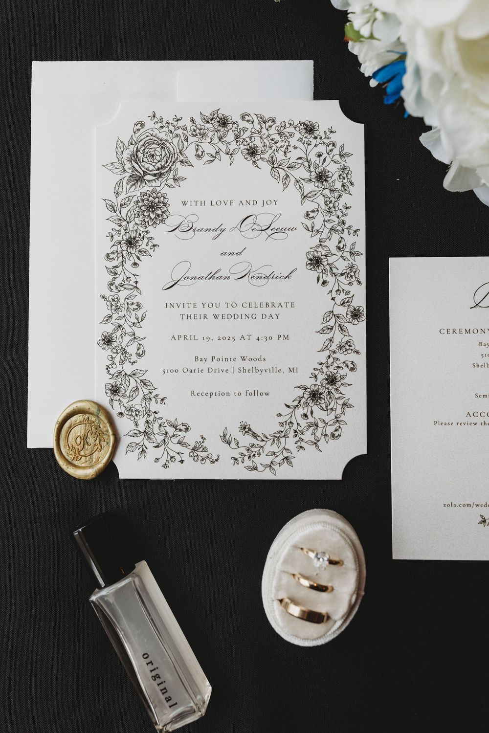 Elegant wedding invitations with delicate floral borders and gold wax seals displayed on dark background with white roses.