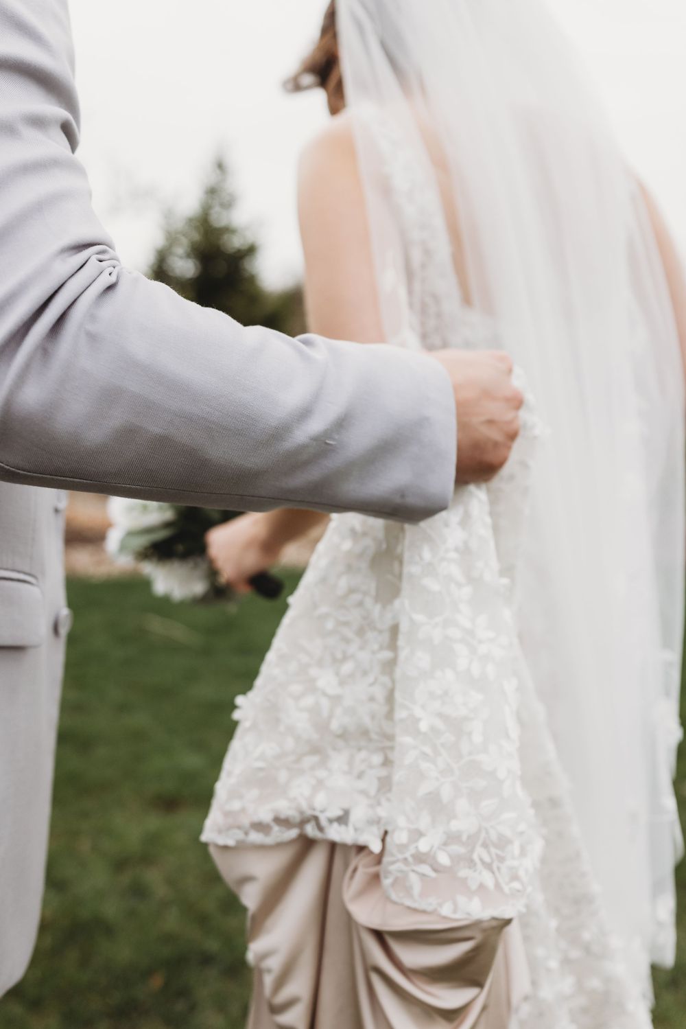 Elegant wedding couple shown from shoulders down wearing gray suit and white lace dress standing outdoors in garden setting.