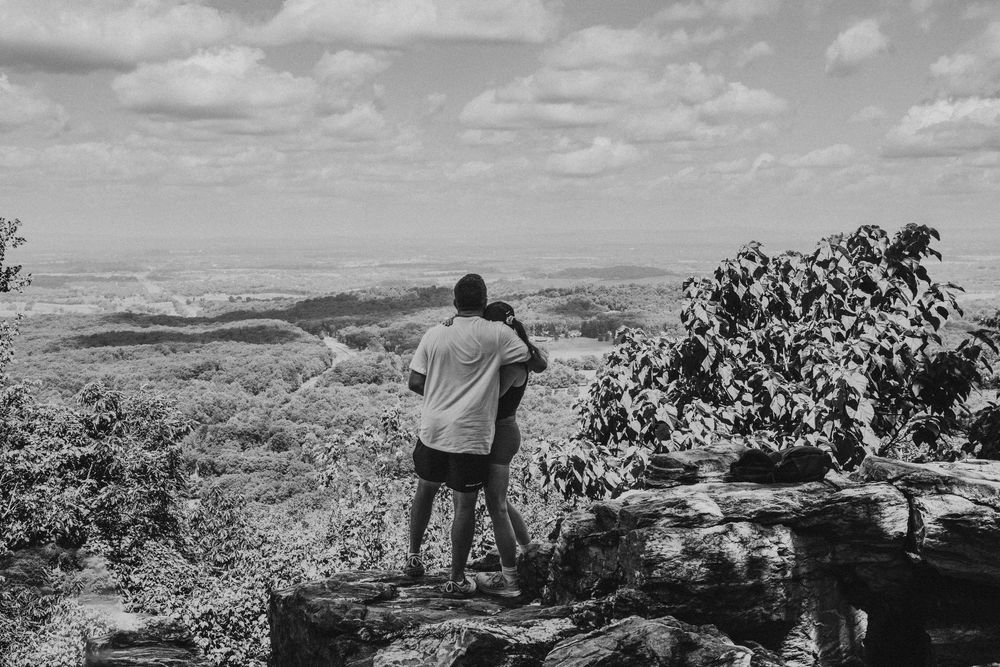 A person stands at a scenic mountain overlook gazing at the vast hilly landscape stretching into the horizon in black and white.