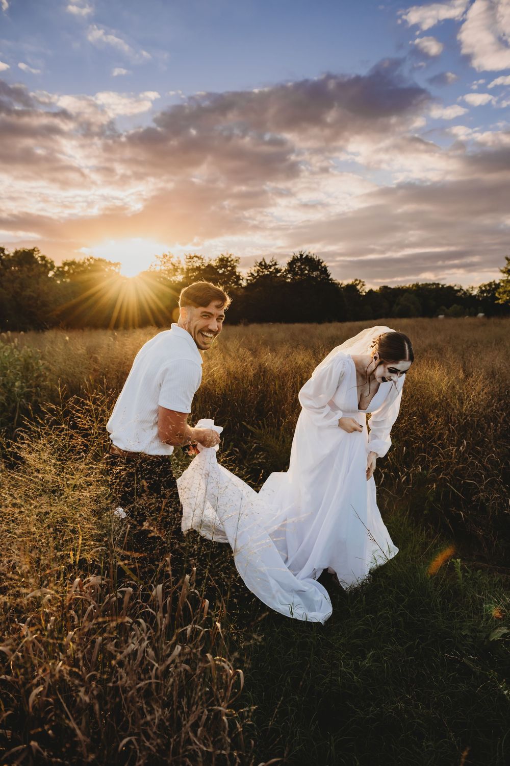 A couple in wedding attire embraces in a golden field at sunset as warm sunlight filters through dramatic clouds.