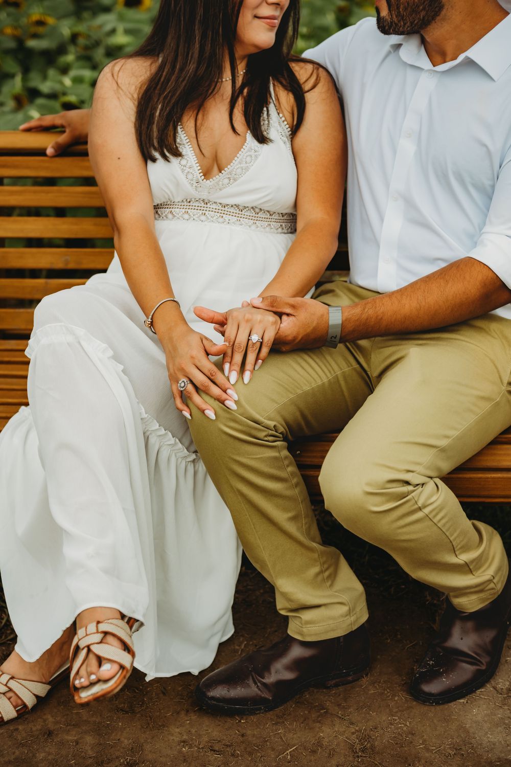 Couple sitting on wooden bench with person wearing white dress and beaded detail showing ring and person in khaki pants.
