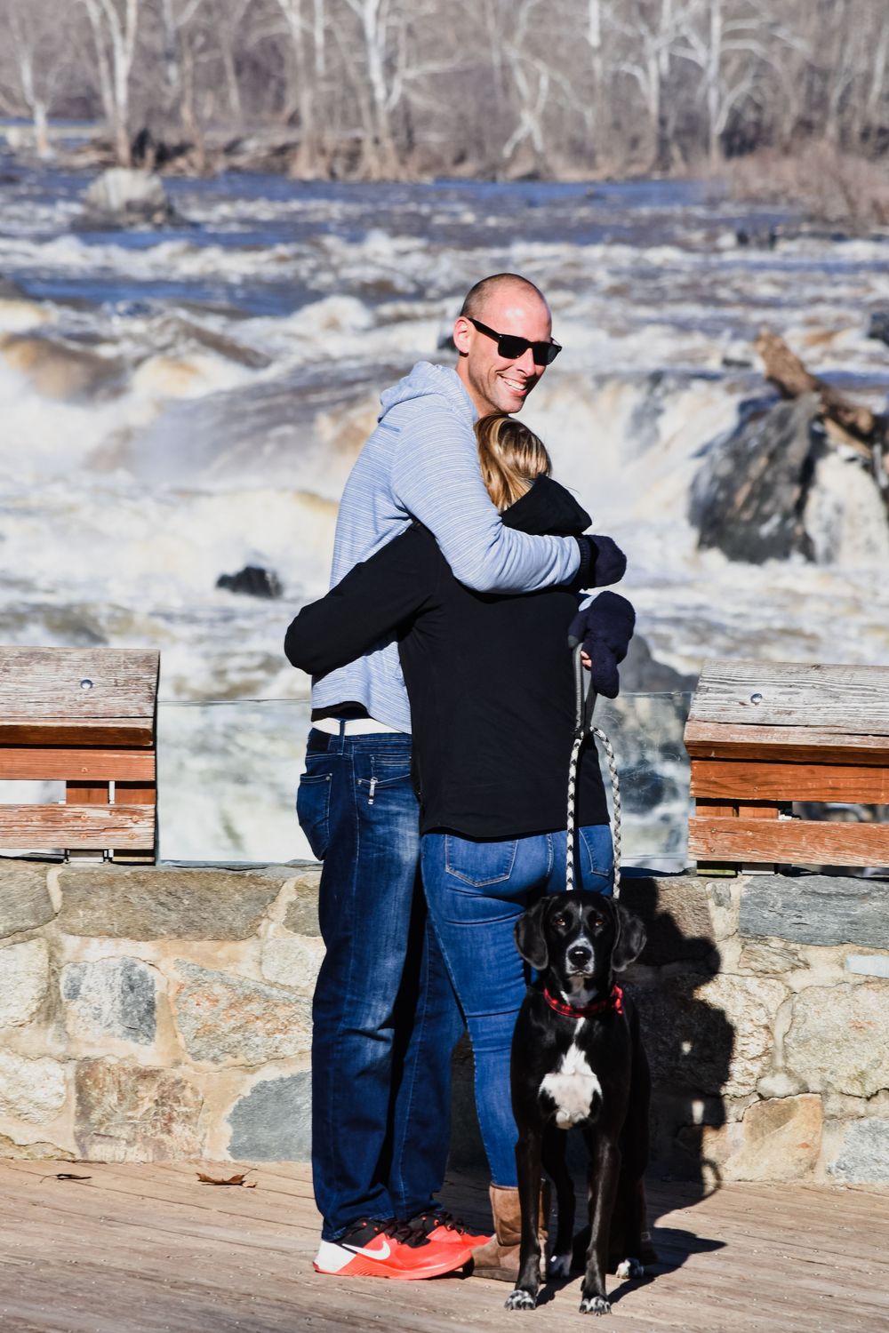 A couple embraces while sightseeing at a rushing waterfall overlook with their black dog on a sunny winter day.
