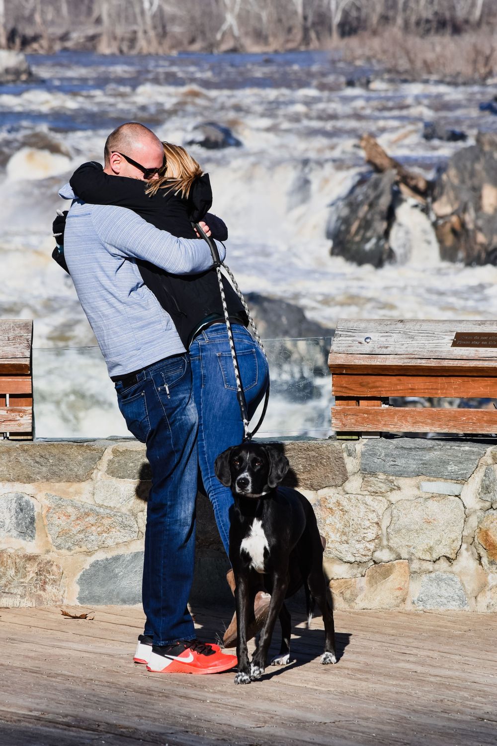 Person kneeling down to embrace a black dog on a wooden deck overlooking a rocky waterfront on a winter day.
