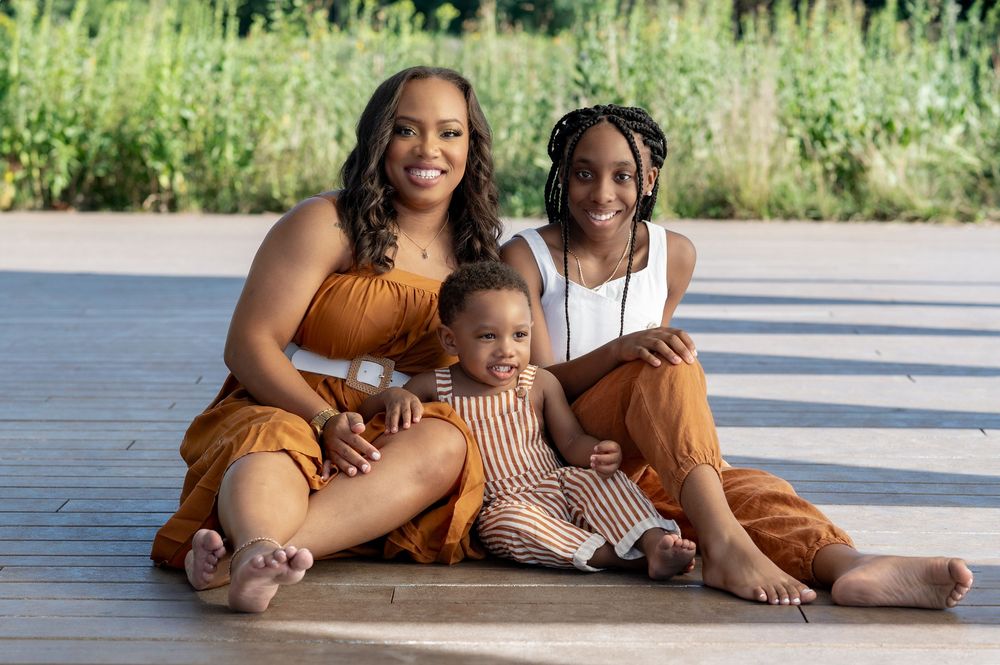 A family wearing coordinated brown and rust-colored outfits sits together on a wooden deck in warm sunlight.