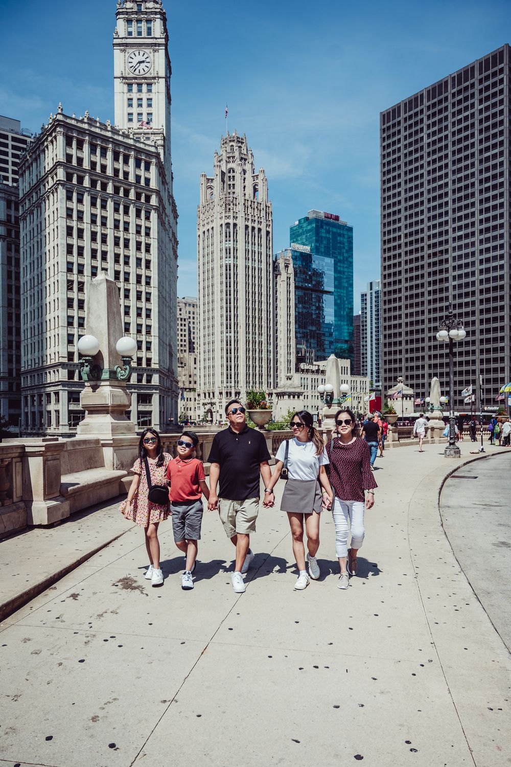 A family walking along the DuSable Bridge in downtown Chicago with iconic architectural buildings in background.